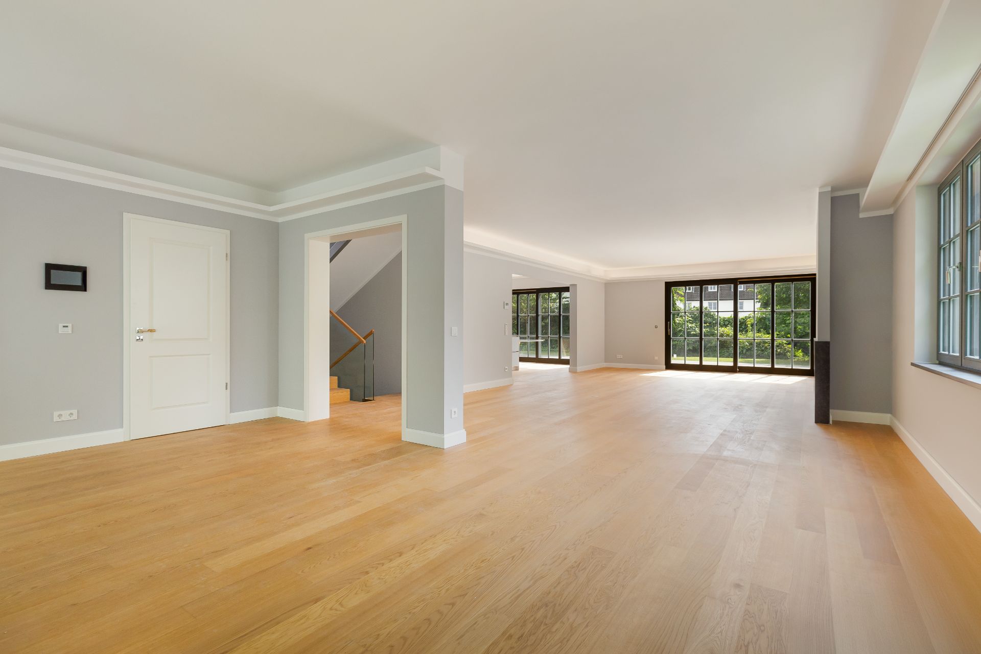 Großes, leeres Wohnzimmer mit Holzfußboden, grauen Wänden und großen Fenstern mit Blick ins Grüne.
