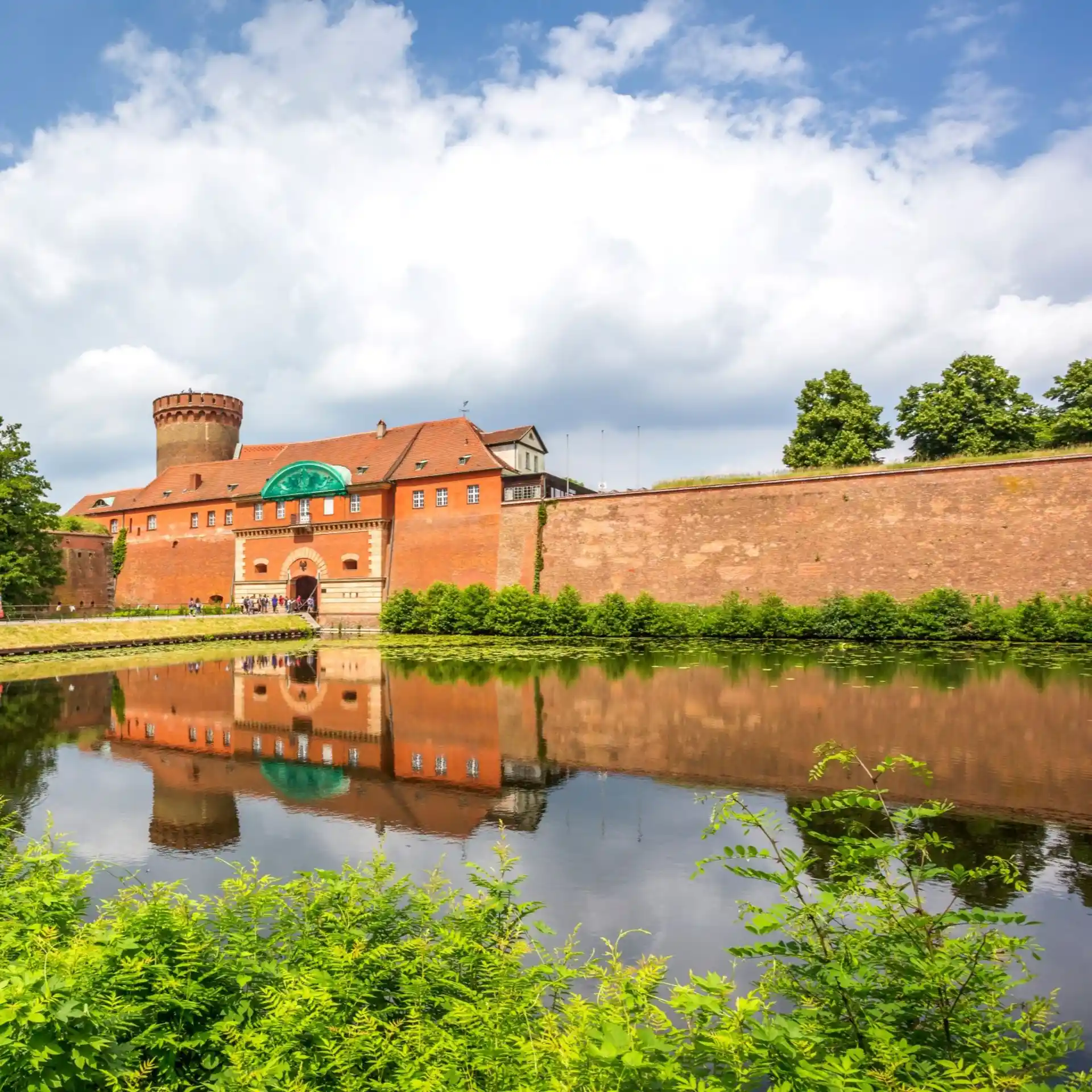Rote Backsteinburg mit Turm, Wassergraben und viel Grün im Vordergrund.