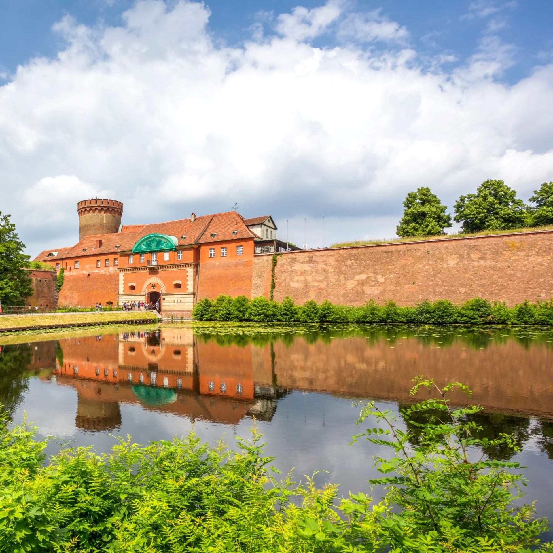 Rote Backsteinburg mit Turm, Wassergraben und viel Grün im Vordergrund.