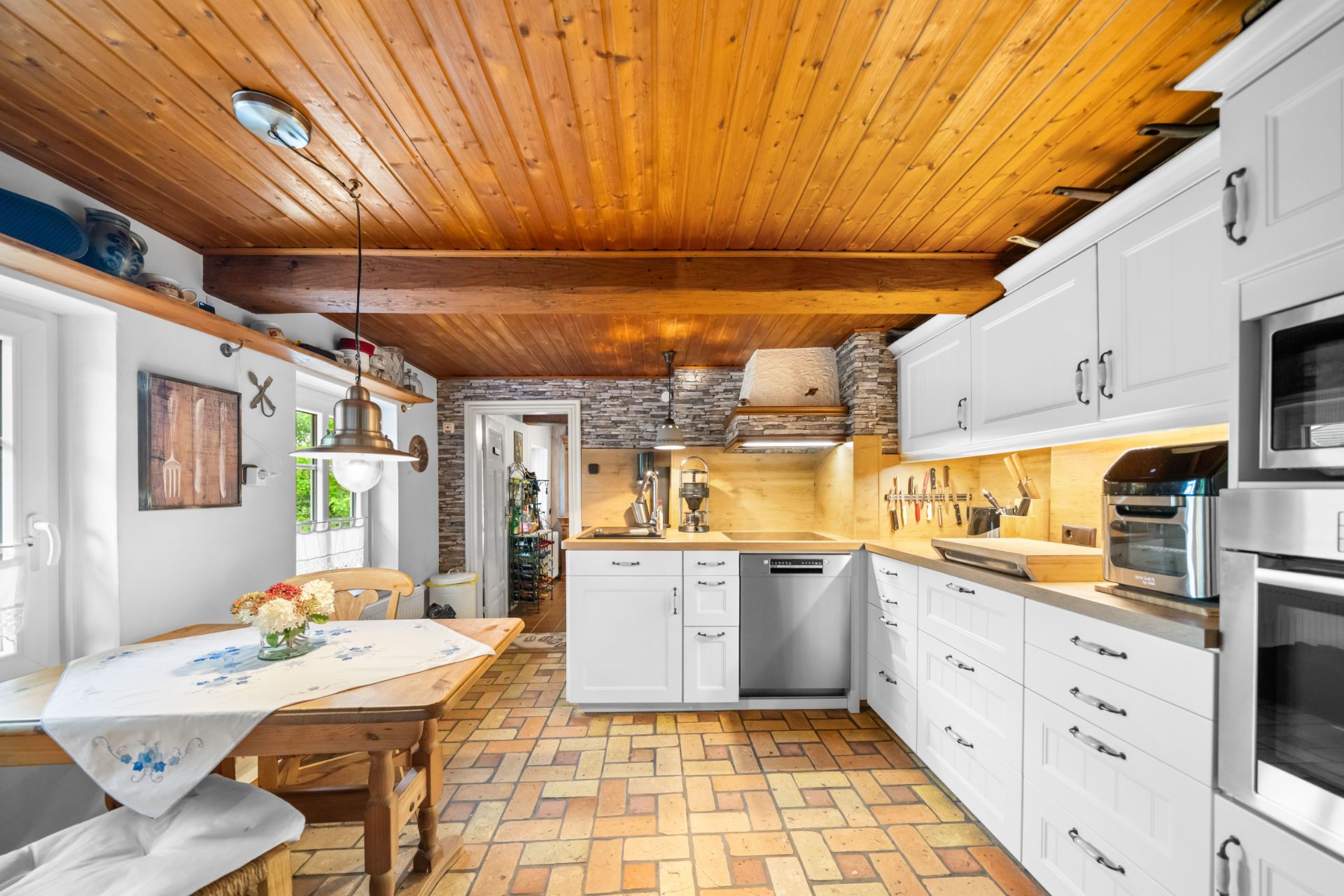 Helle Küche mit weißen Schränken, Holzbalkendecke und kleinem Esstisch mit Blumen.