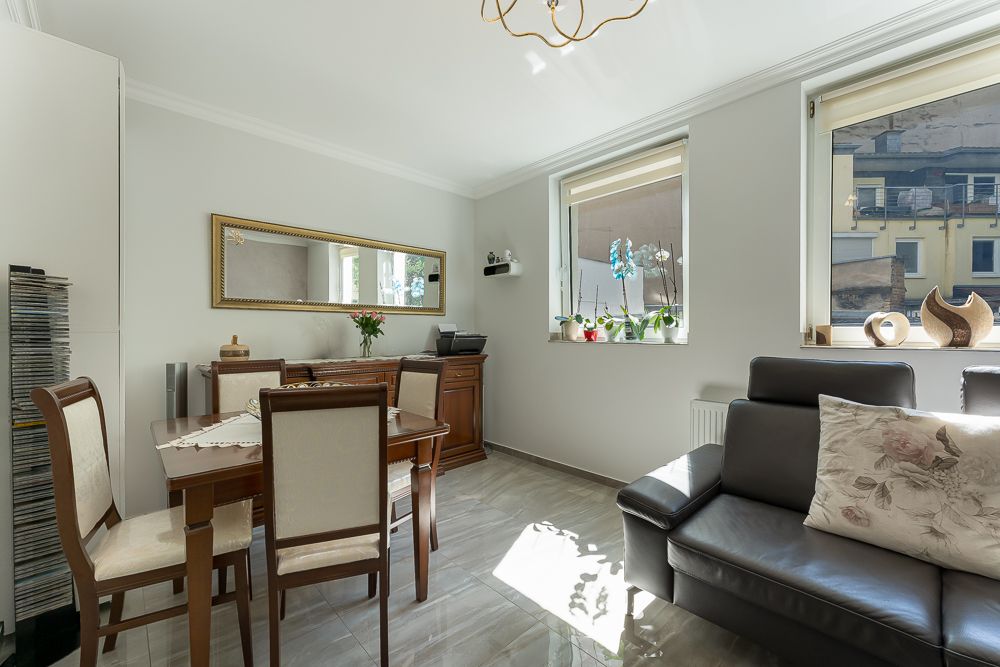 Helles Wohnzimmer mit Esstisch, vier Stühlen, Sofa und zwei großen Fenstern mit Pflanzen.