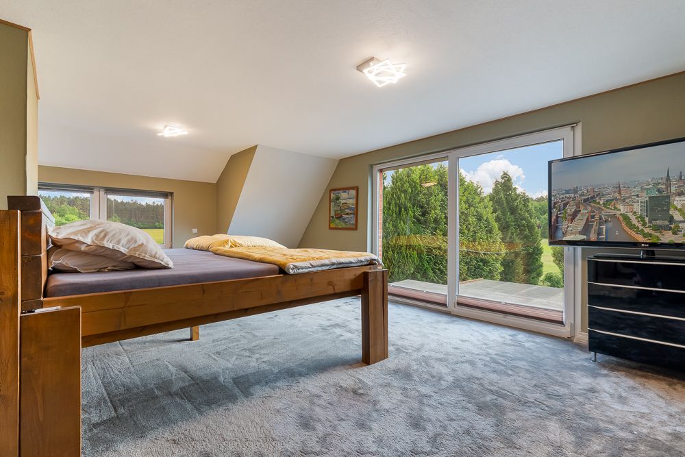 Schlafzimmer mit großem Bett, Teppichboden, Fernseher und Zugang zu einem Balkon mit Blick ins Grüne.