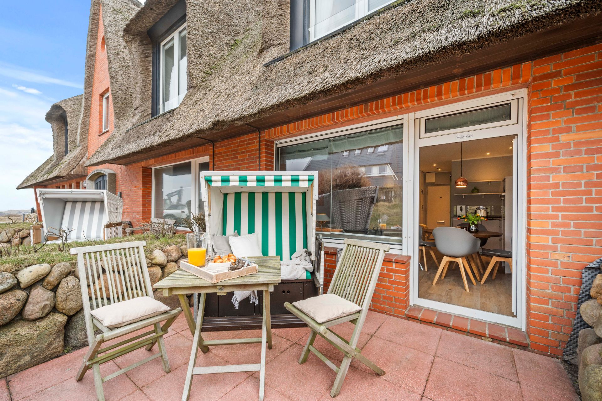 Gemütliche Terrasse mit zwei Stühlen, Strandkorb und gedecktem Tisch vor einem Backsteinhaus.