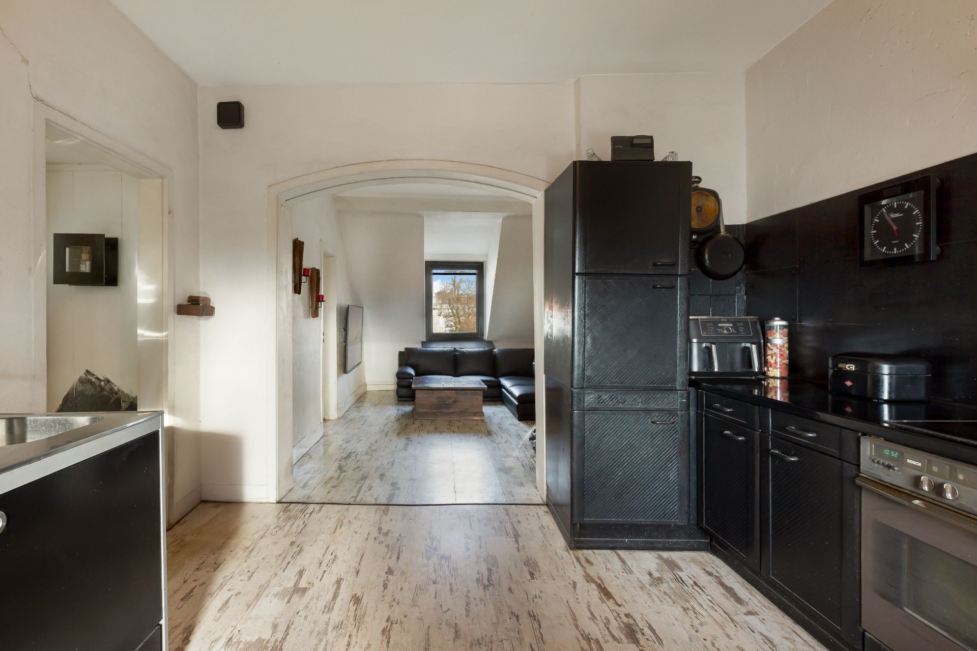 Blick aus einer modernen schwarzen Küche in ein Wohnzimmer mit Sofa und Fenster.
