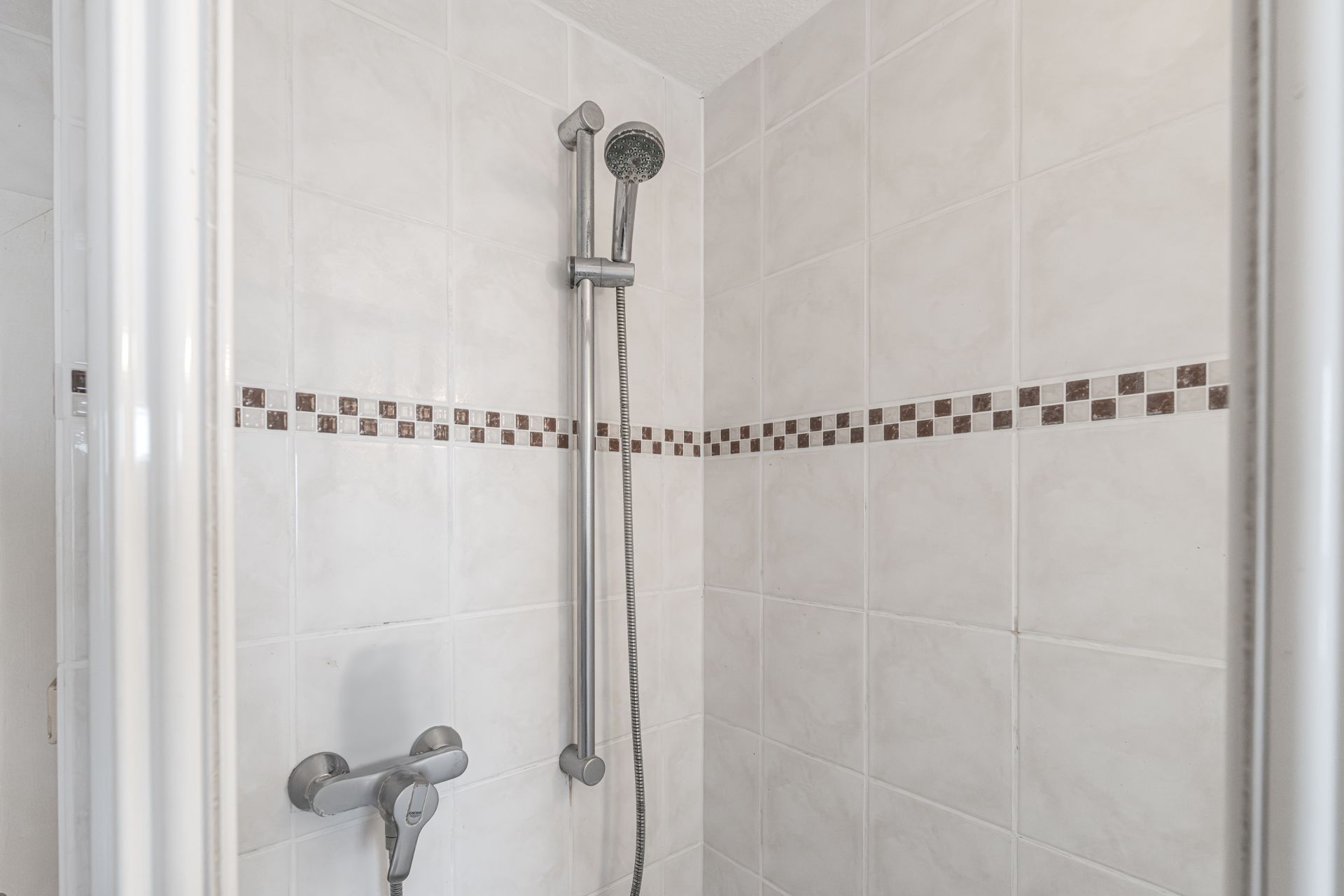 Nahaufnahme einer Dusche mit Handbrause und weißen Fliesen, mittig braune Zierleiste.