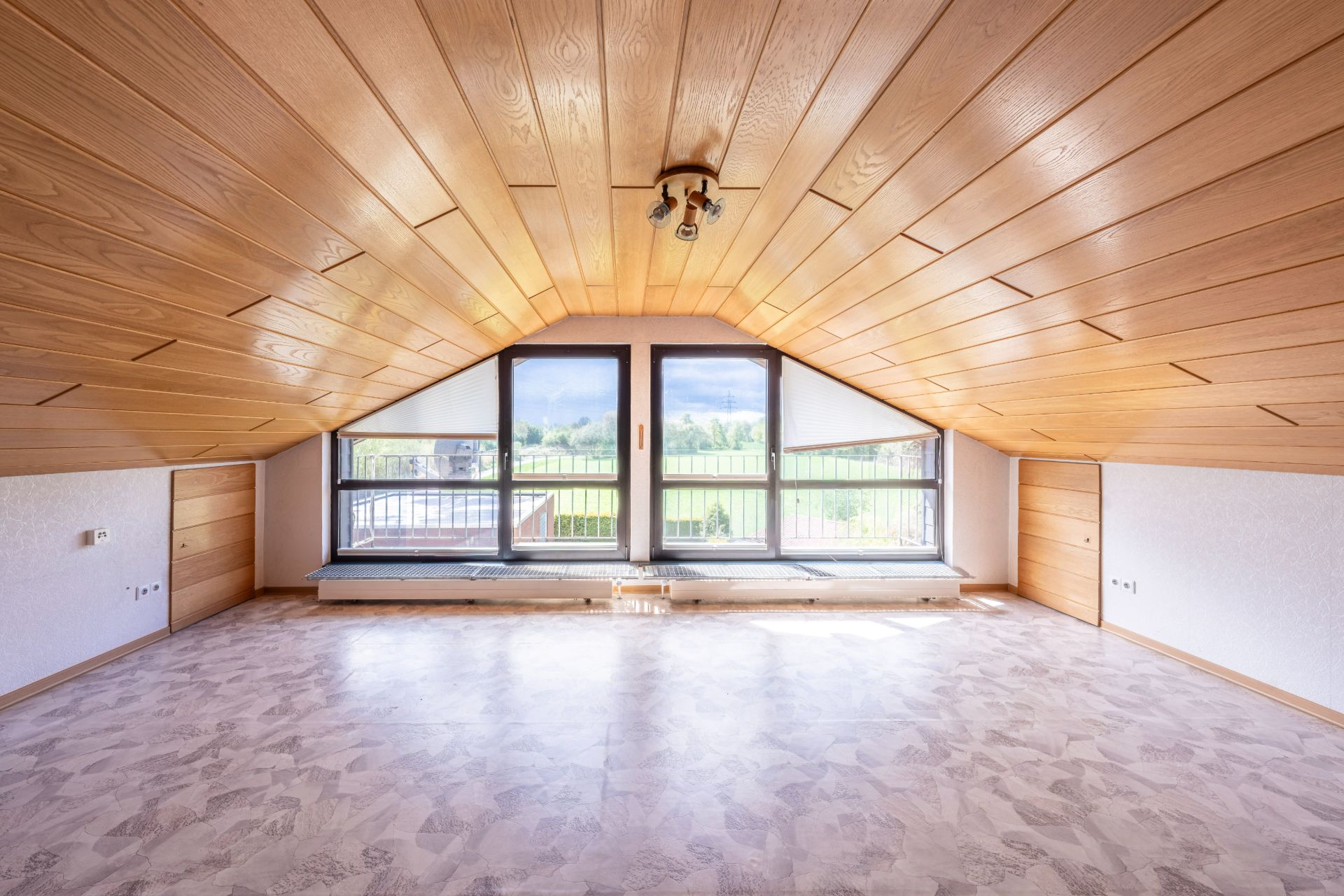 Leerer Dachbodenraum mit Holzdecke, großen Fenstern und Ausblick ins Grüne.