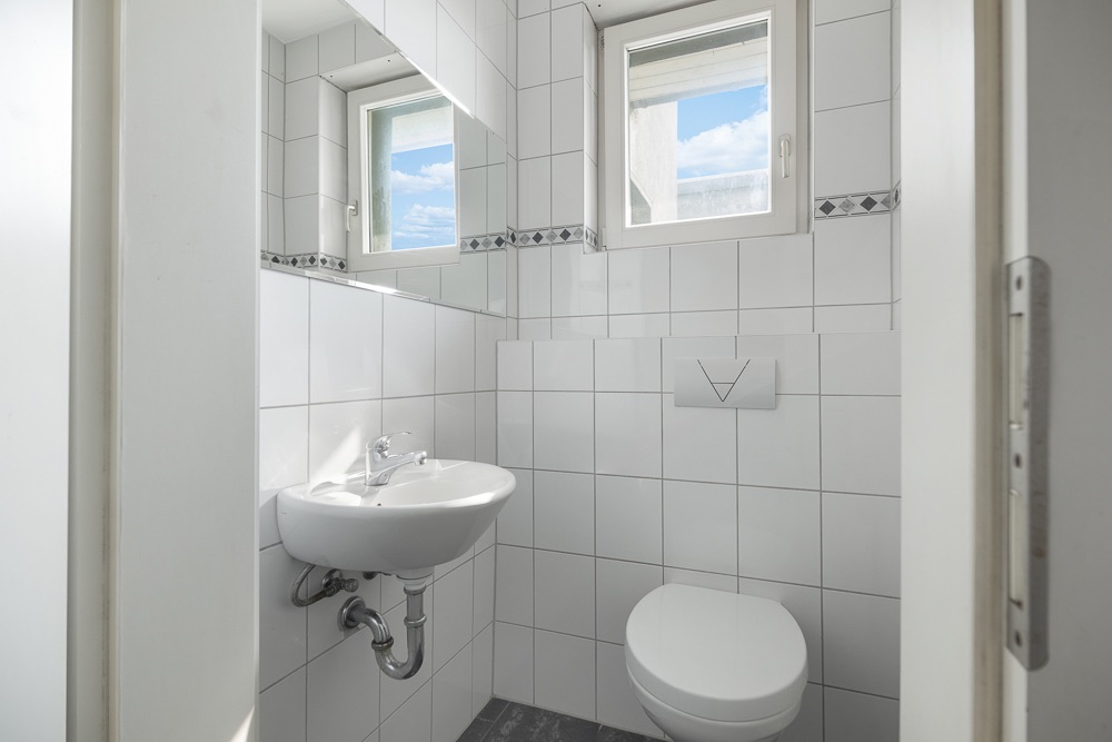 Kleines, weiß gefliestes Badezimmer mit Wand-WC, Waschbecken und zwei Fenstern.