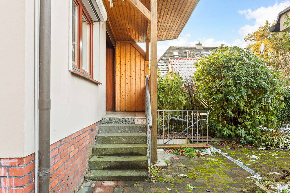 Außentreppe mit Geländer führt zu einer Eingangstür; rechts Büsche und gepflasterter Weg.