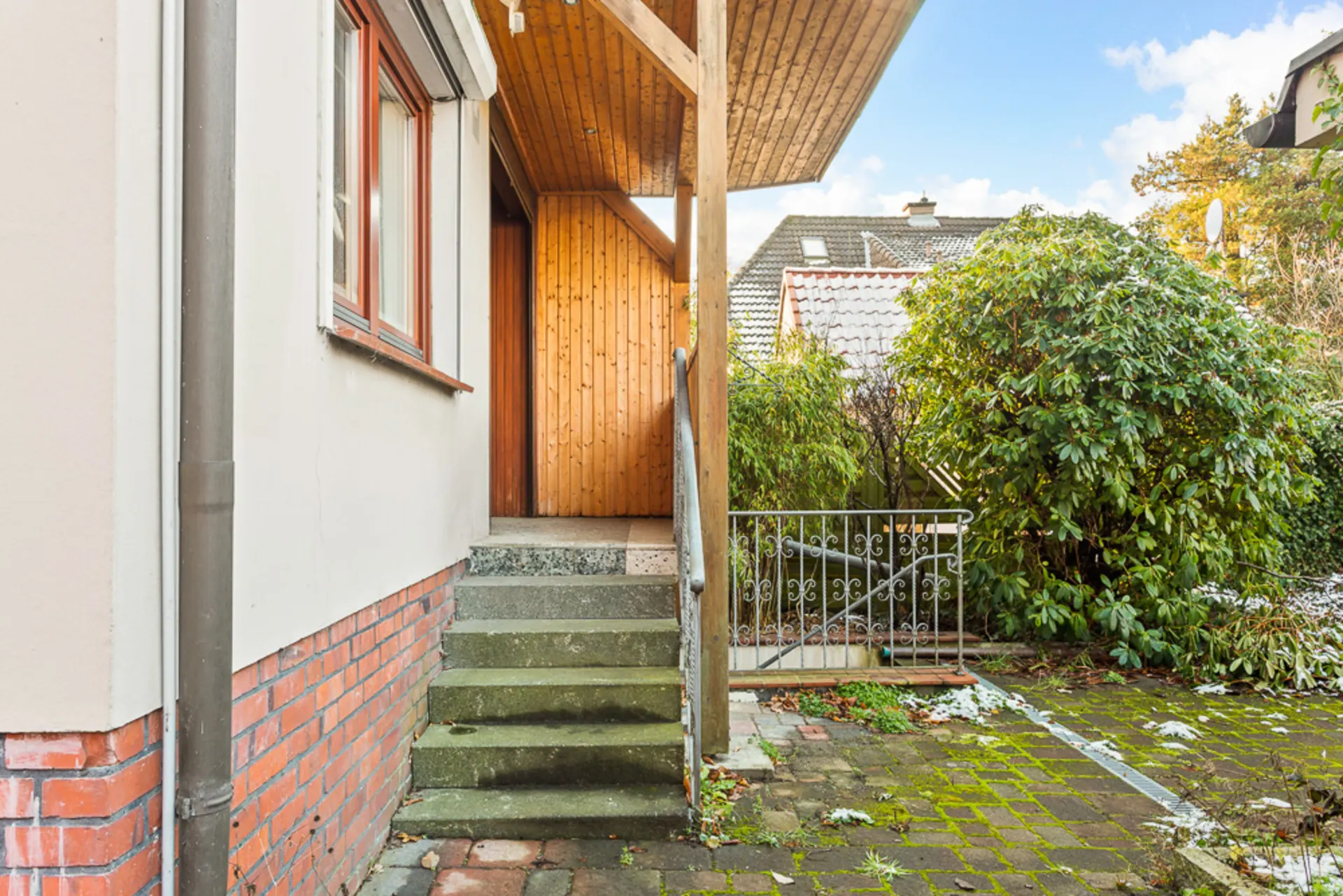Außentreppe mit Geländer führt zu einer Eingangstür; rechts Büsche und gepflasterter Weg.