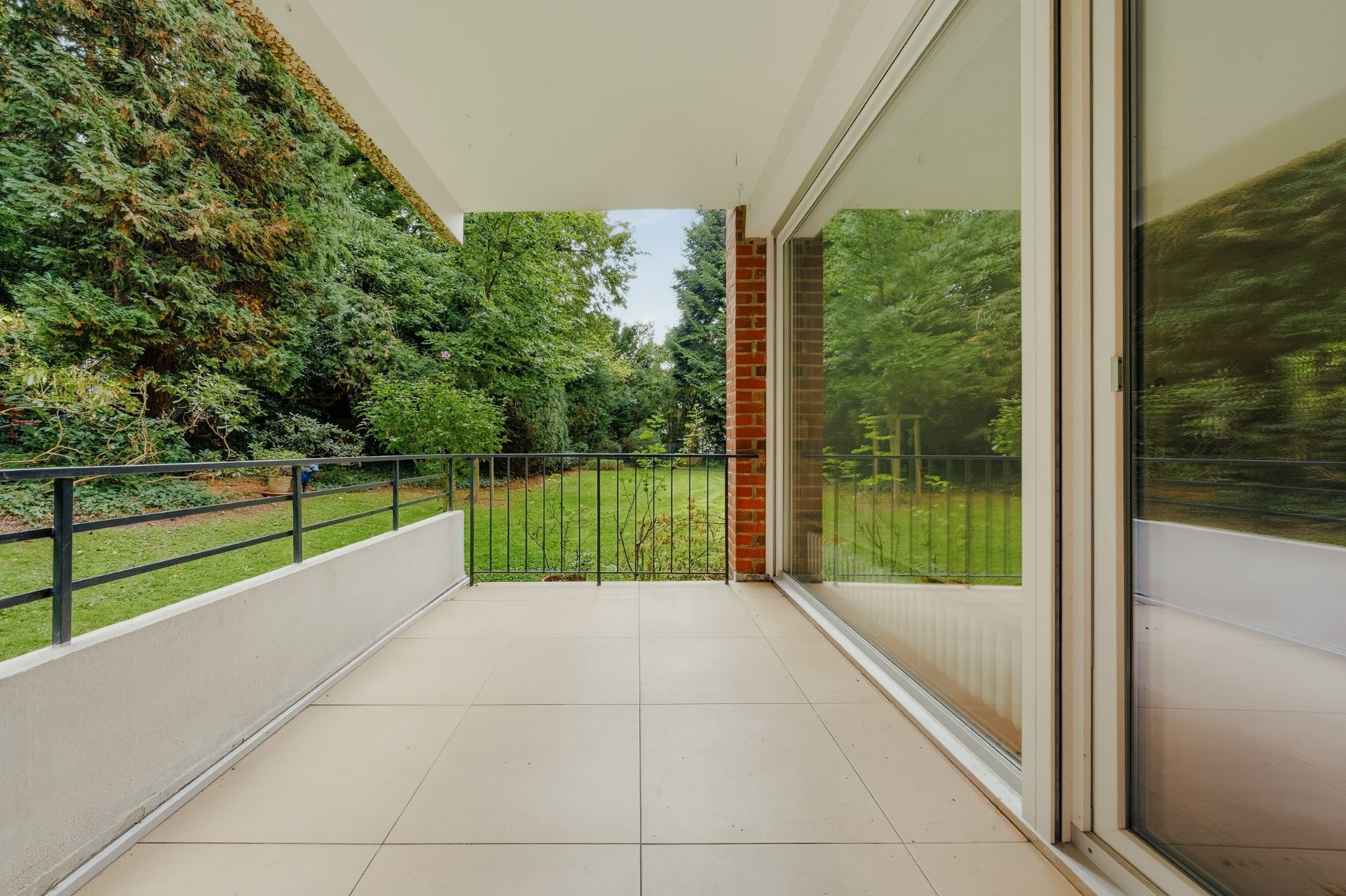 Überdachter Balkon mit Fliesenboden und Blick auf einen Garten mit viel Grün.