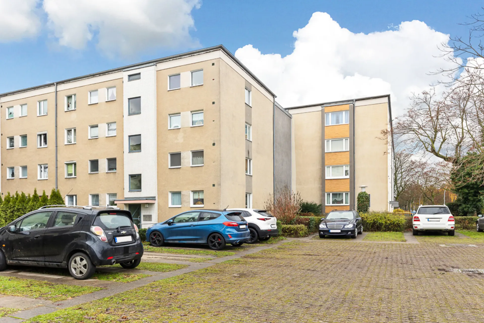 Mehrfamilienhaus mit mehreren geparkten Autos auf einem Parkplatz davor.