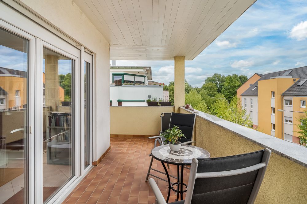 Überdachter Balkon mit zwei schwarzen Stühlen, rundem Tisch und Pflanze, Blick auf Wohngebäude.