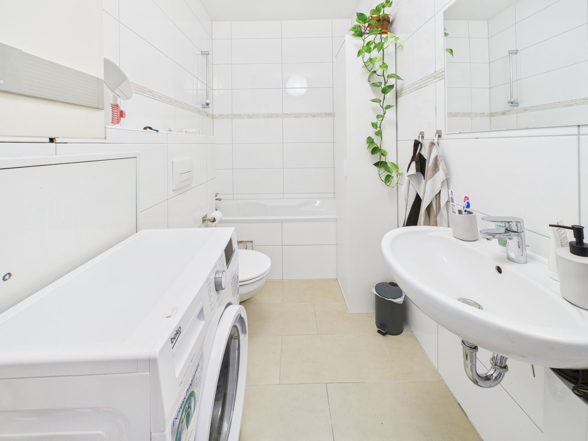 Helles Badezimmer mit Waschmaschine, Waschbecken, Badewanne, Toilette und Pflanzen an der Wand.