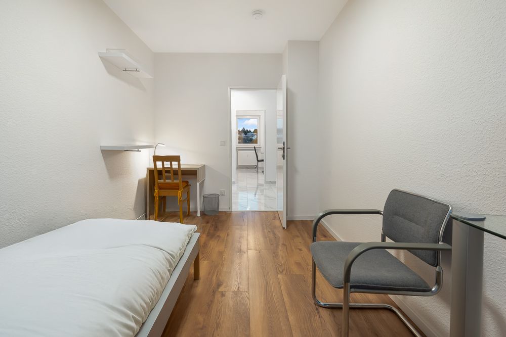 Ein schlicht eingerichtetes Schlafzimmer mit Einzelbett, Schreibtisch, Stuhl und Sessel auf Holzfußboden.