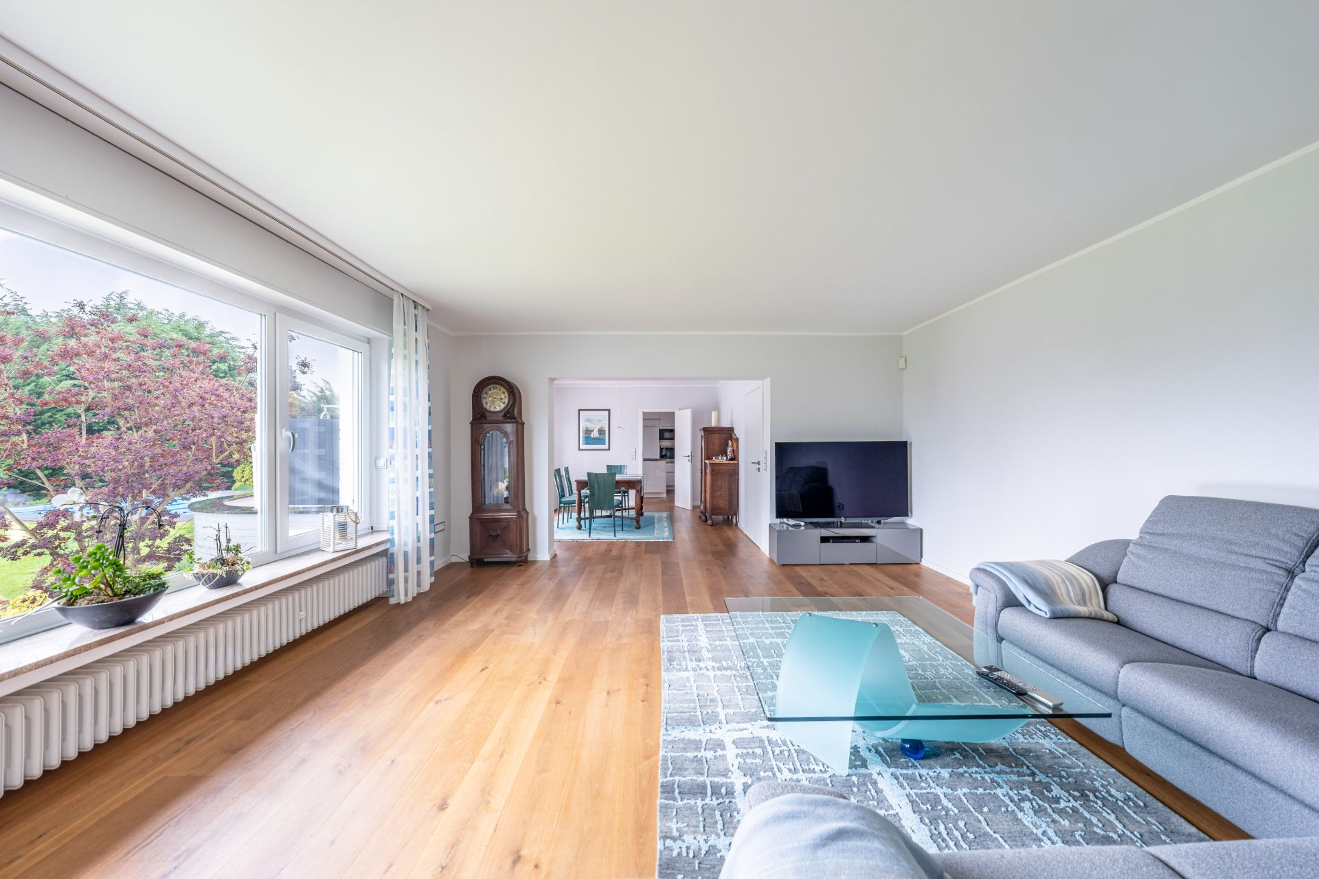 Helles Wohnzimmer mit großen Fenstern, grauem Sofa, Glastisch und Fernseher, Blick in Esszimmer.