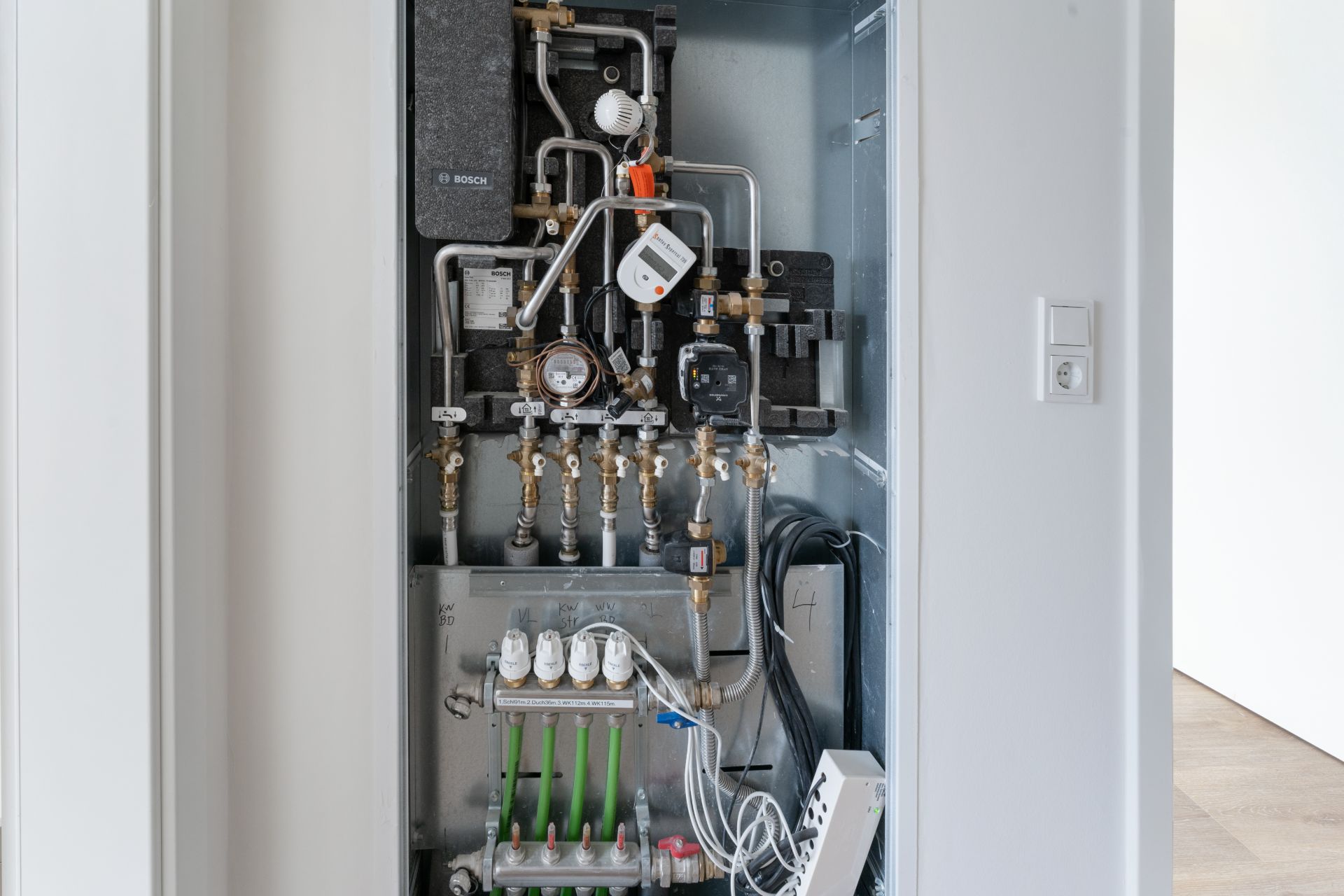 Geöffneter Technikschrank mit Rohren, Ventilen und Messgeräten in einer Wohnung.