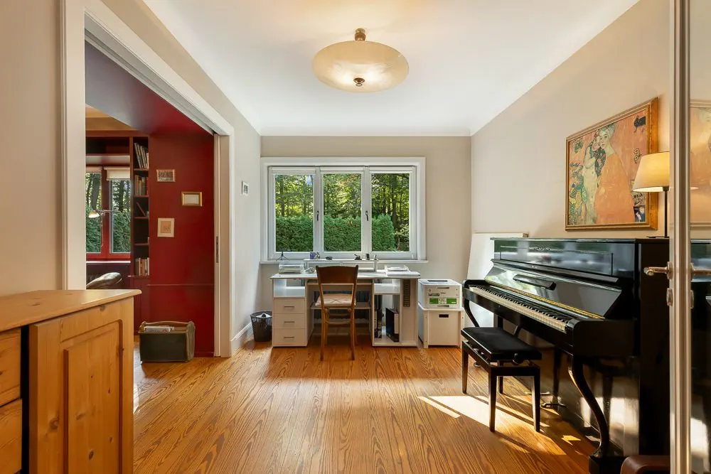 Helles Zimmer mit Schreibtisch am Fenster und schwarzem Klavier an der Seitenwand.