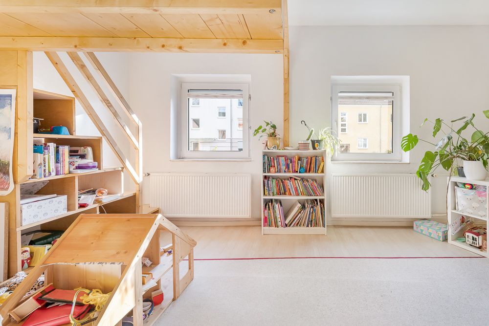 Helles Kinderzimmer mit zwei Fenstern, Bücherregalen, Pflanzen und einem Holzspielhaus.