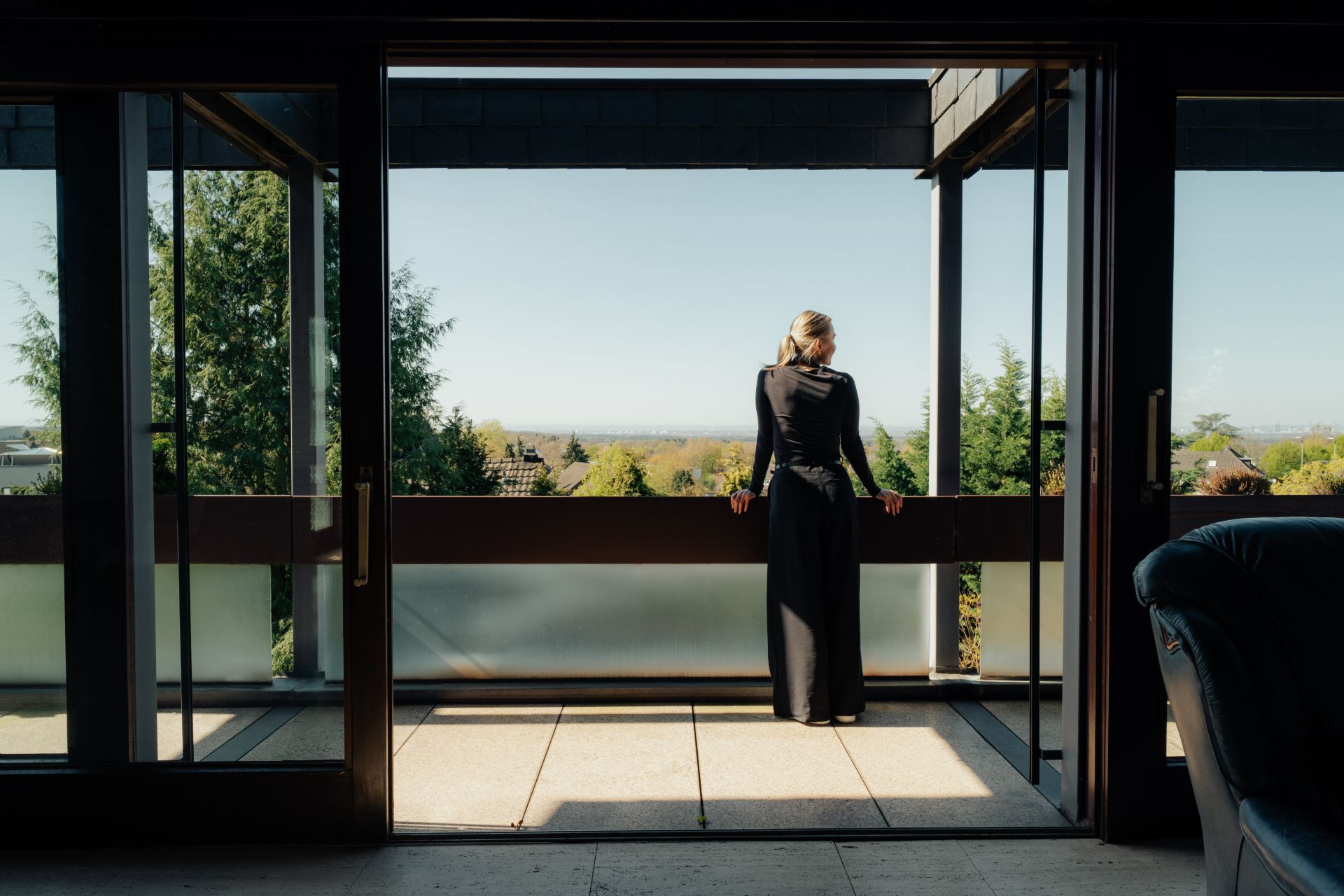 Frau steht auf einem Balkon und blickt auf Bäume und den Horizont.