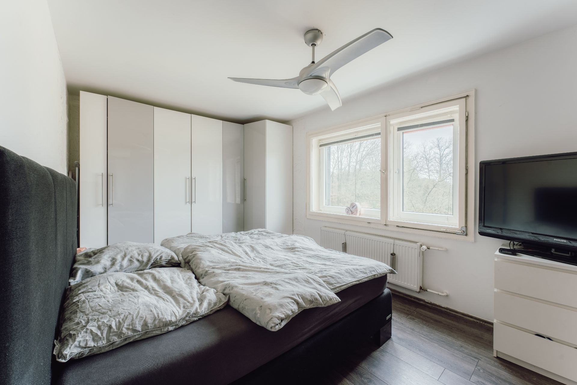 Modernes Schlafzimmer mit Bett, großem Schrank, Fernseher, Deckenventilator und zwei Fenstern.