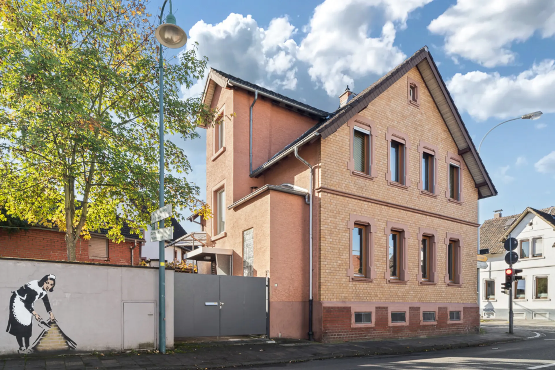 Zweistöckiges Wohnhaus aus Backstein an einer Straßenecke, Wandmalerei von Frau mit Besen links.