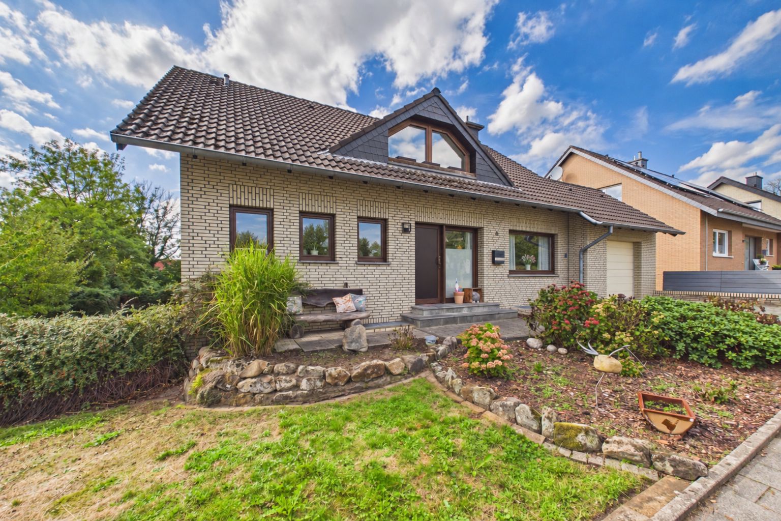 Einfamilienhaus mit Vorgarten, Steintreppe und Blumenbeeten, unter blauem Himmel.
