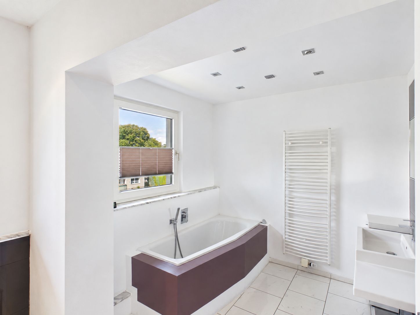Helles Badezimmer mit Badewanne, Fenster, Handtuchheizkörper und kleinem Waschbecken.