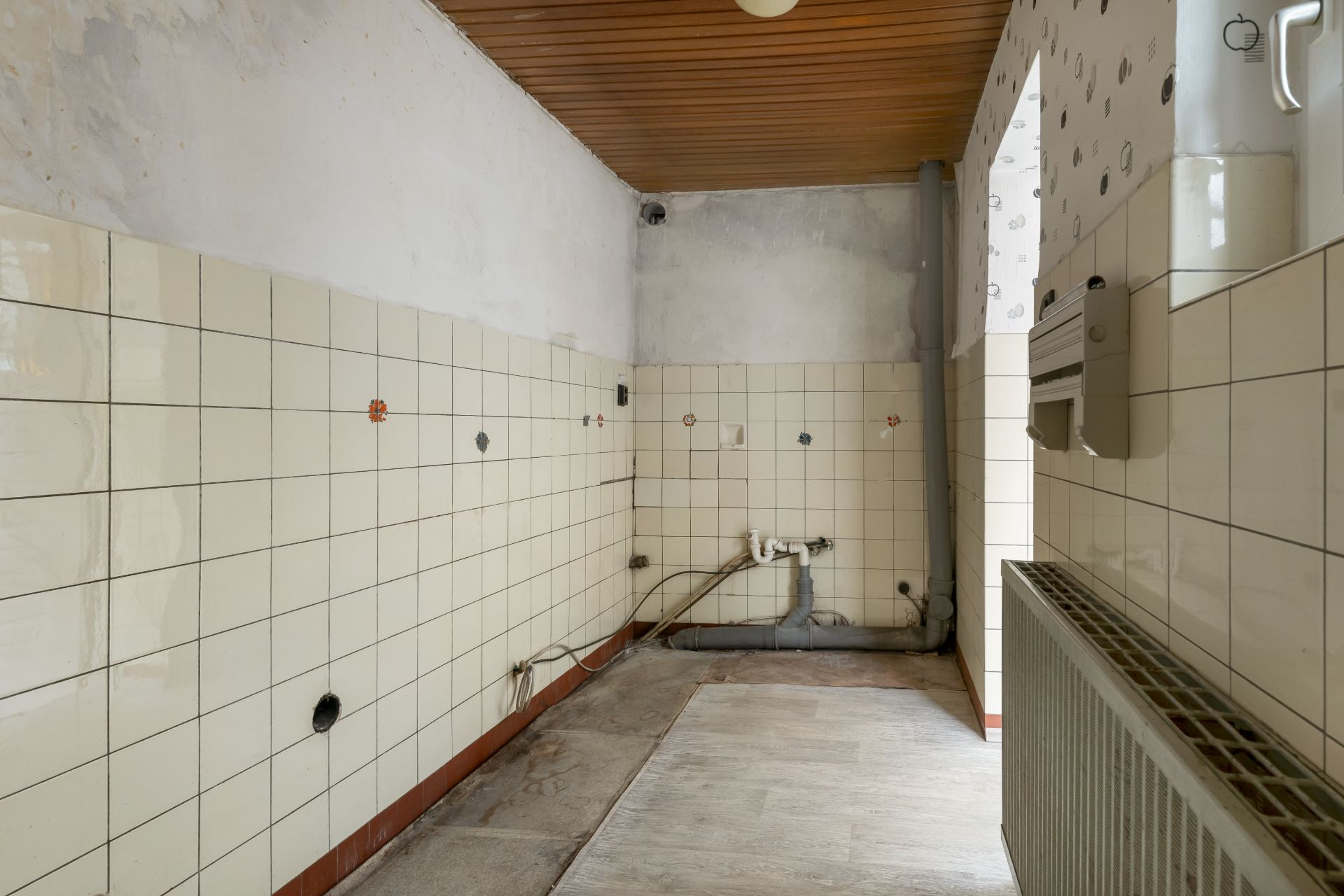 Leerer Raum mit weißen Fliesen, sichtbaren Rohren und einem Heizkörper an der Wand.