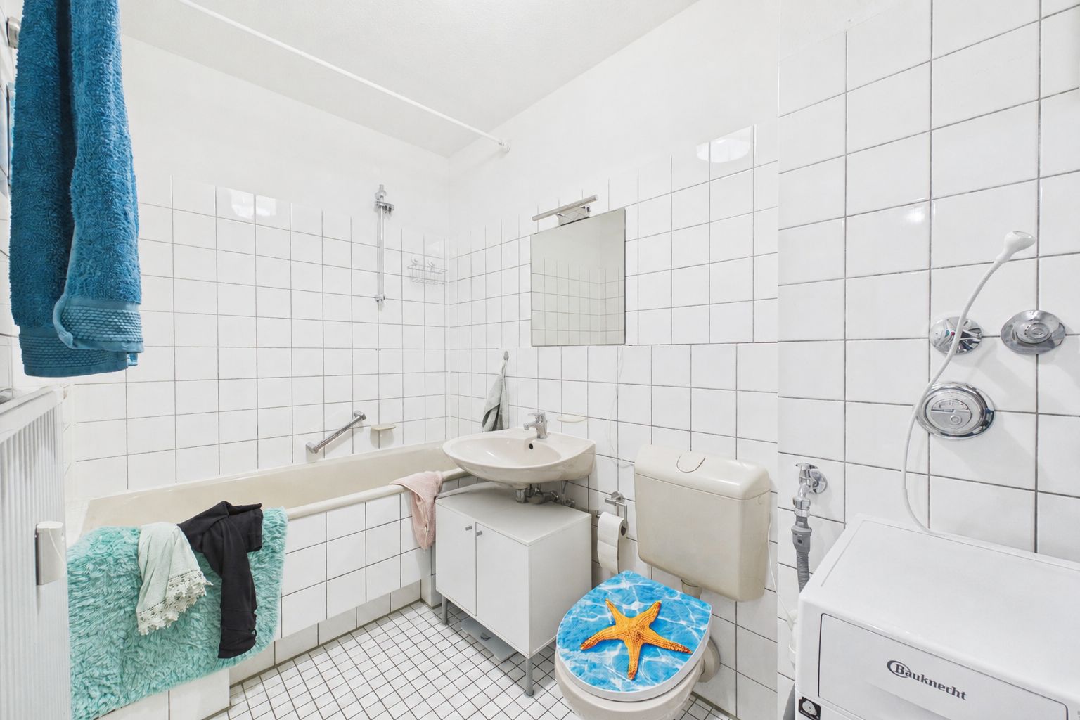 Weiß gefliestes Badezimmer mit Badewanne, Waschbecken, Toilette und Waschmaschine.
