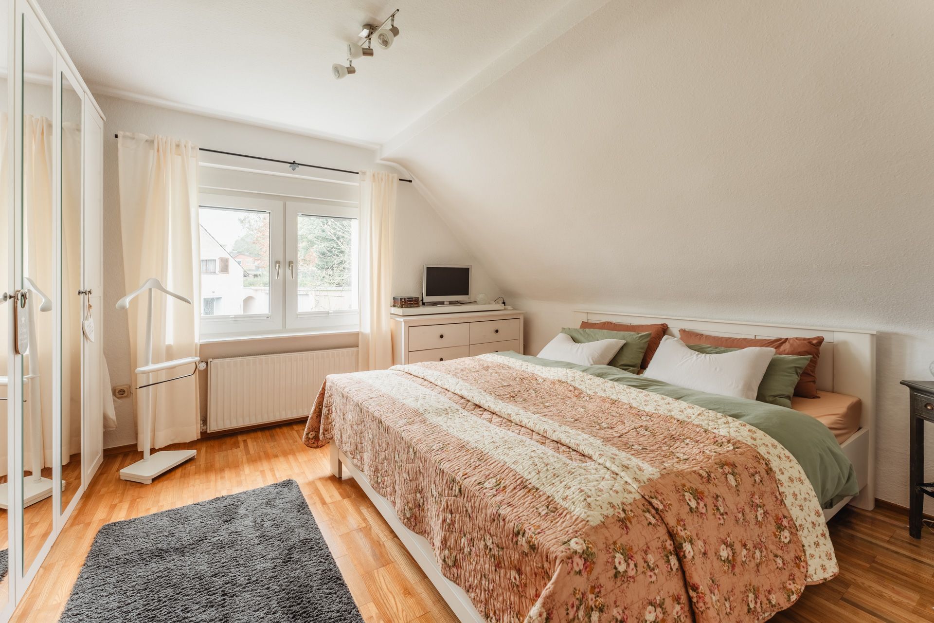 Helles Schlafzimmer mit Doppelbett, Kommode, Teppich und großem Fenster mit Vorhängen.