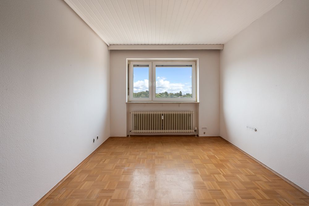 Leeres Zimmer mit Parkettboden, weißer Decke und Fenster mit Heizkörper darunter.
