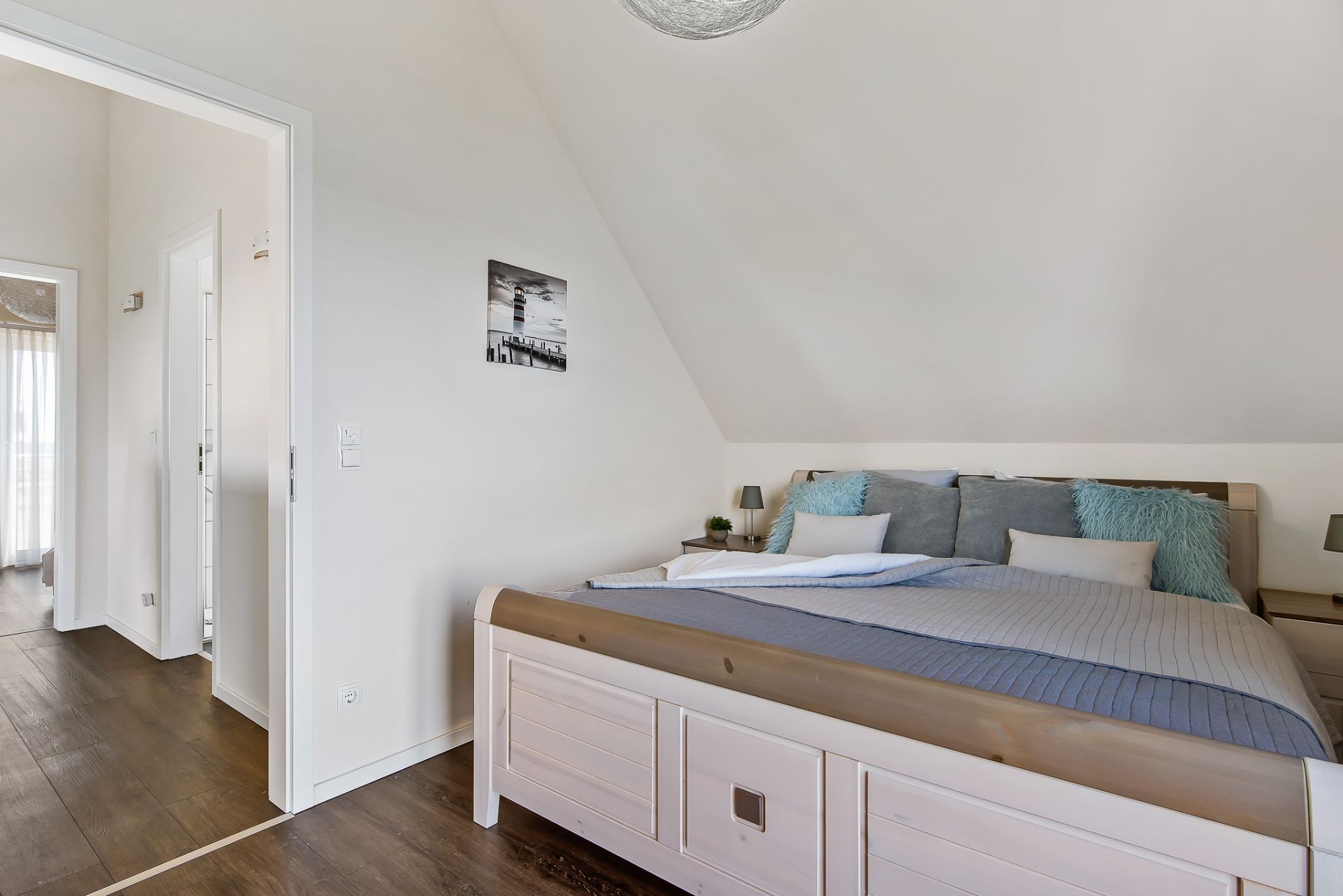 Schlafzimmer mit großem Bett, weißen Wänden und Holzboden, Blick in einen Flur.