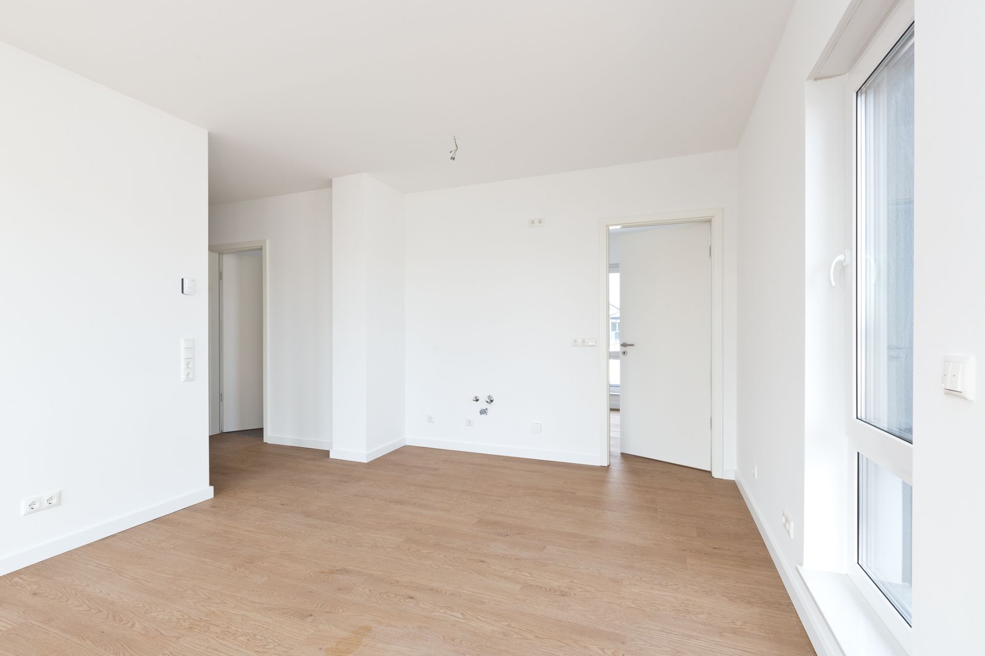 Heller, leerer Raum mit weiß gestrichenen Wänden und Holzfußboden, große Fenster rechts.
