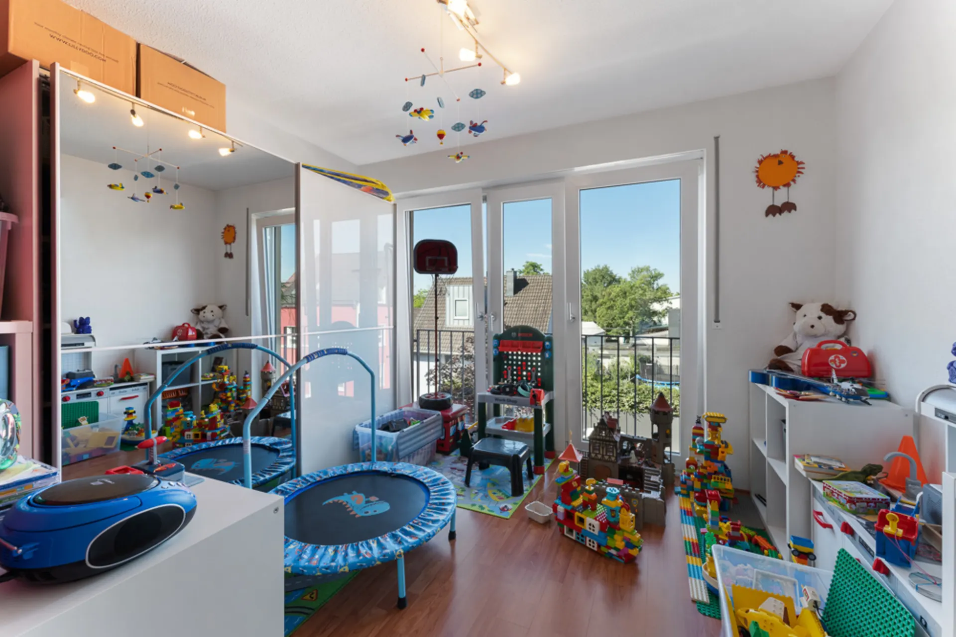 Helles Kinderzimmer mit Spielzeug, Bauklötzen, Trampolin und großen Fenstern zum Balkon.