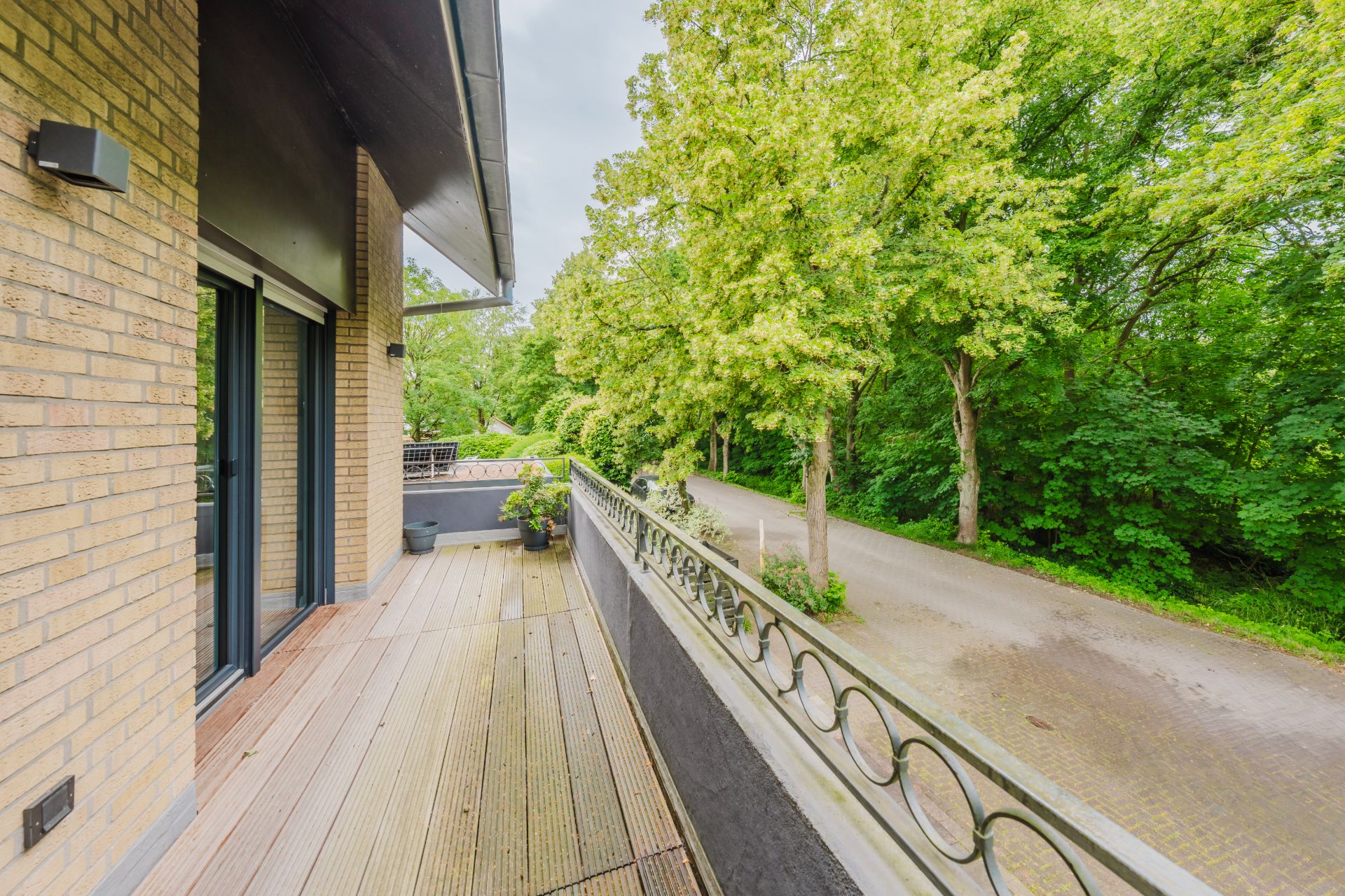 Balkon mit Holzboden, Geländer und Blick auf eine Baumallee neben einer Straße.