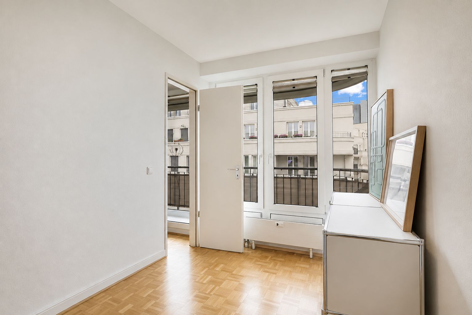 Heller Raum mit Parkettboden, großen Fenstern, offener Tür und Kommoden an der rechten Wand.
