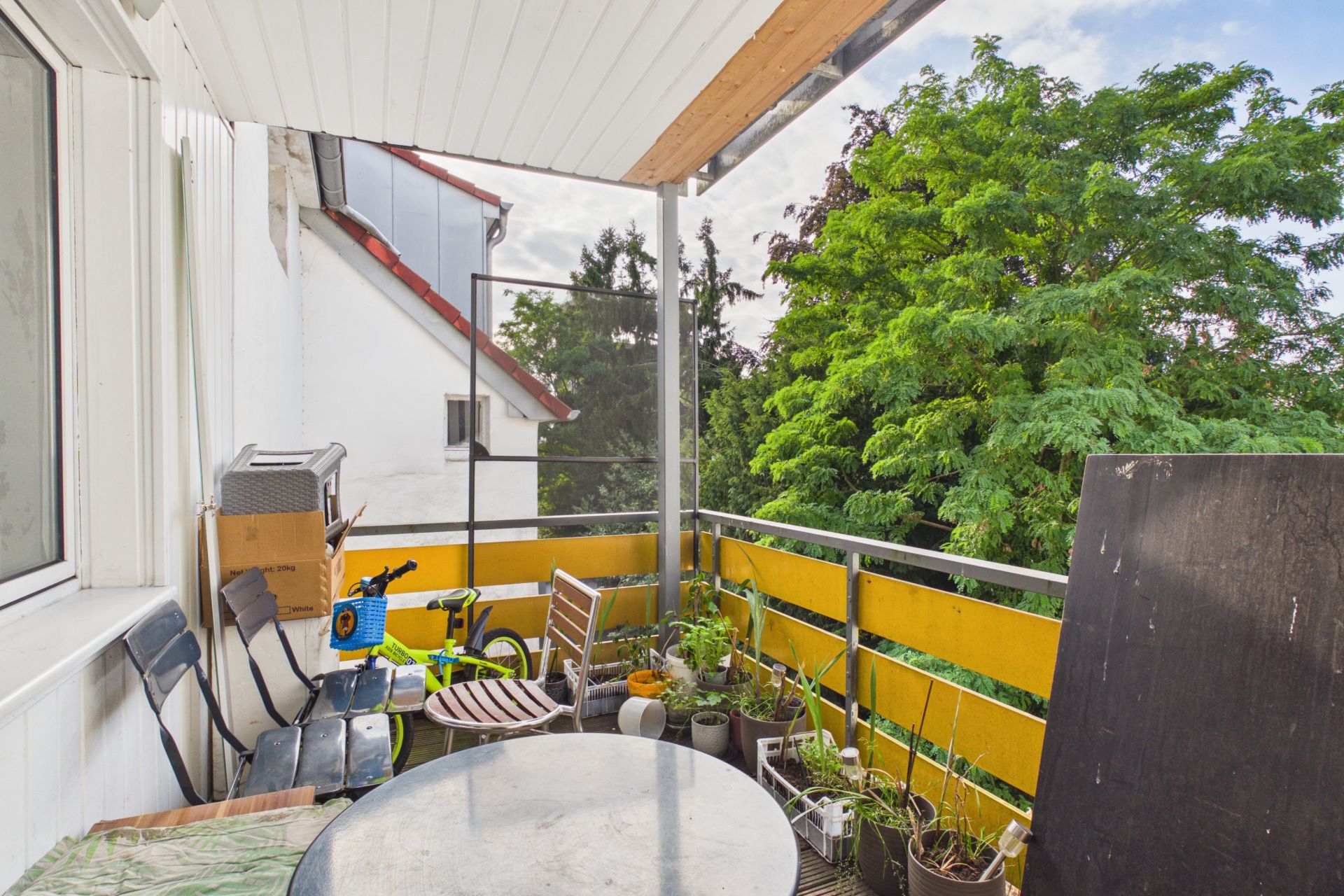 Balkon mit Tisch, Stühlen, Pflanzen und etwas Kinderspielzeug, daneben grüne Bäume.
