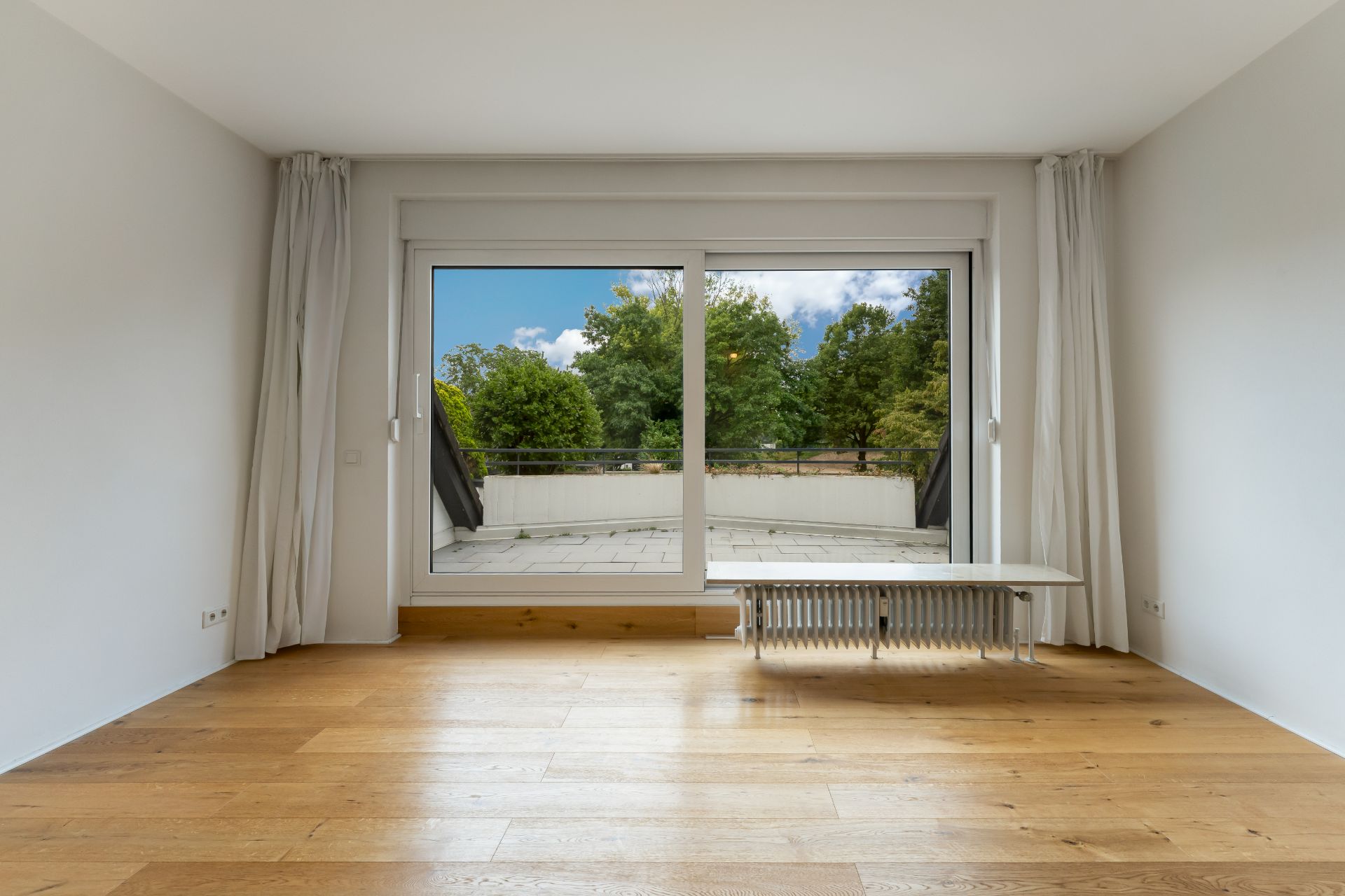 Leerer Raum mit Holzboden, großem Fenster und Blick auf einen Balkon mit Bäumen im Hintergrund.