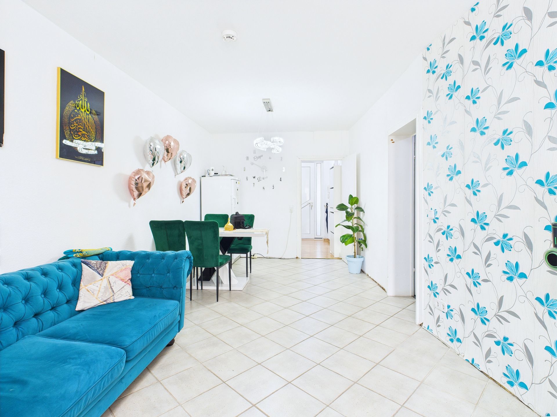 Helles Wohnzimmer mit blauem Sofa, Esstisch, grünen Stühlen und einer Wand mit Blumenmuster.