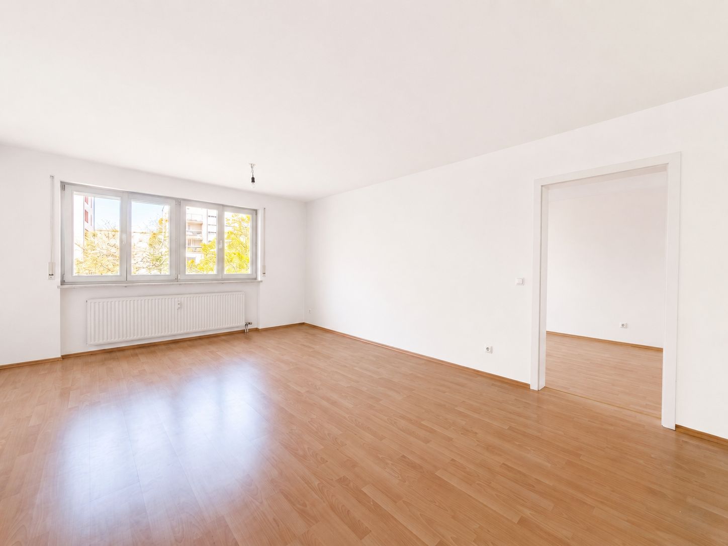 Helles, leeres Zimmer mit Holzfußboden, großen Fenstern und angrenzendem Raum.