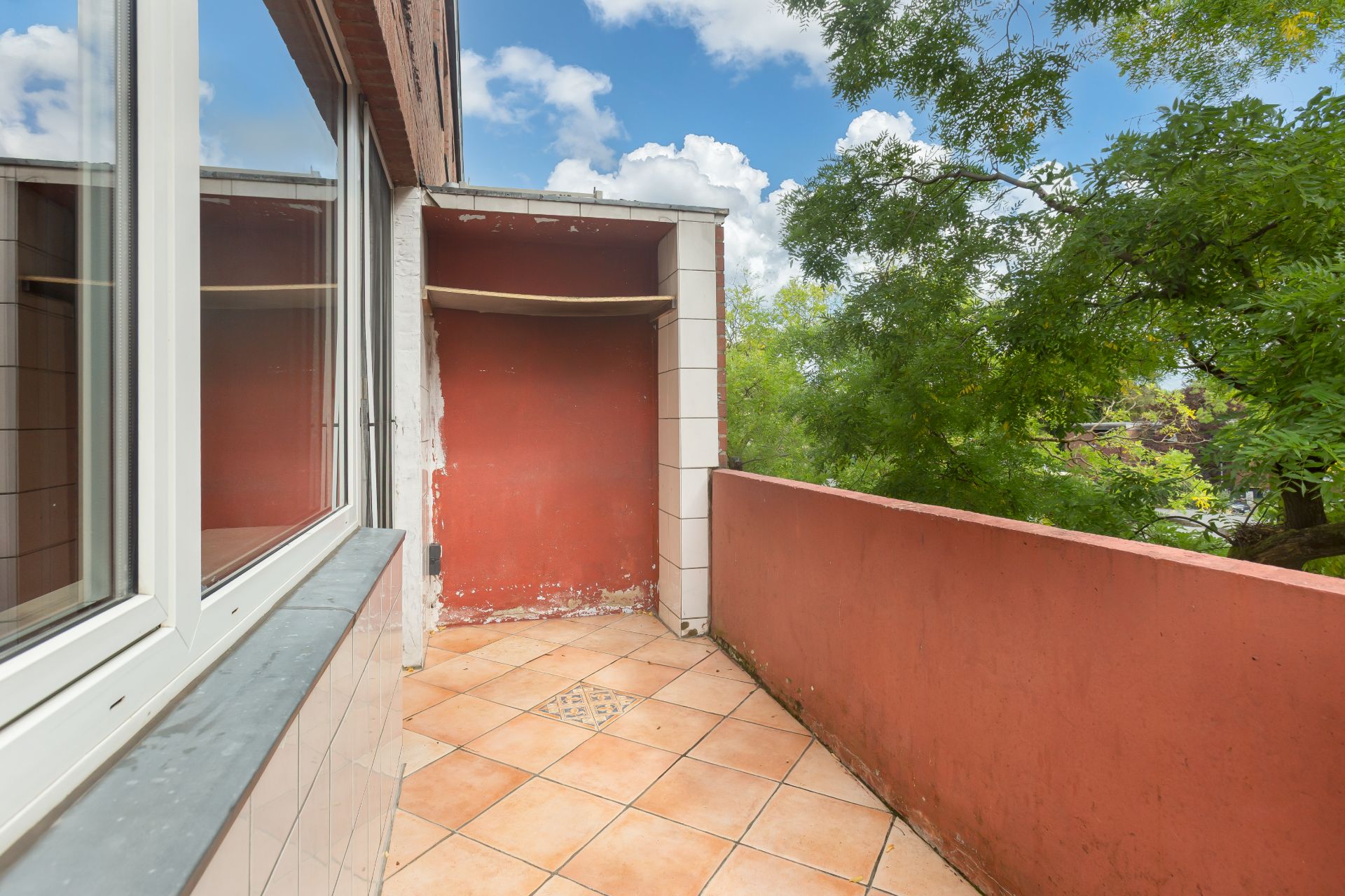 Balkon mit roten Wänden, Fliesenboden und Blick auf grüne Bäume.