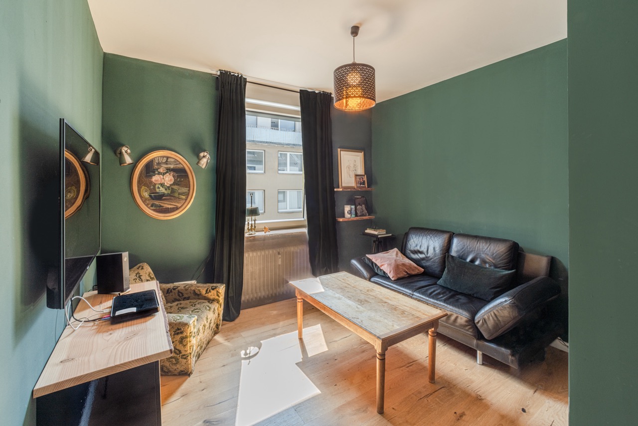 Kleines Wohnzimmer mit grünen Wänden, schwarzem Sofa, Holztisch und Sessel, Fenster mit dunklen Vorhängen.