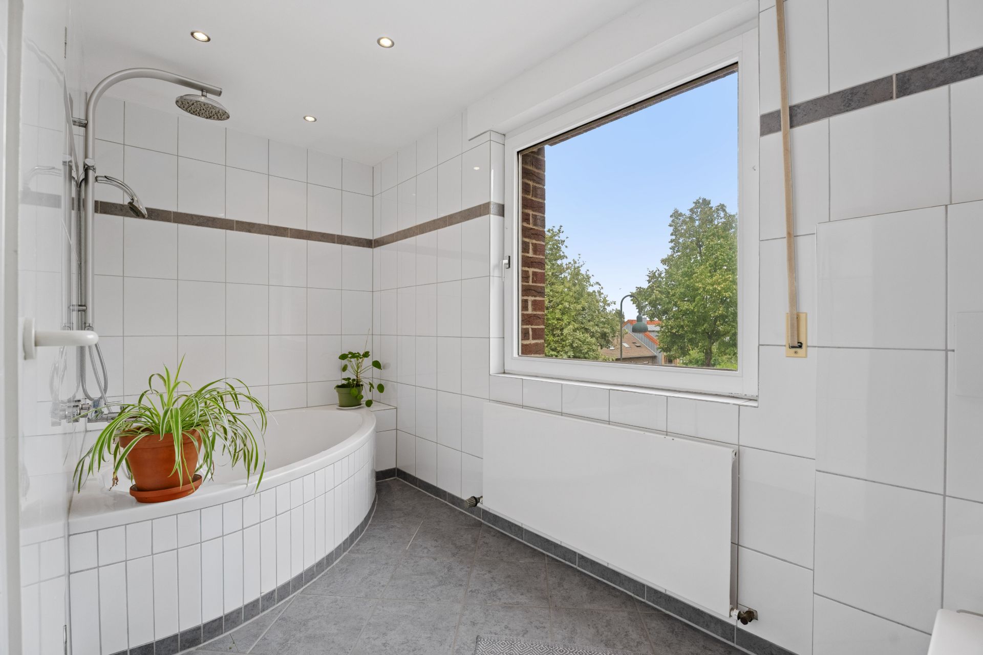 Helles Badezimmer mit Eckbadewanne, weißen Fliesen, Fenster und zwei grünen Pflanzen.