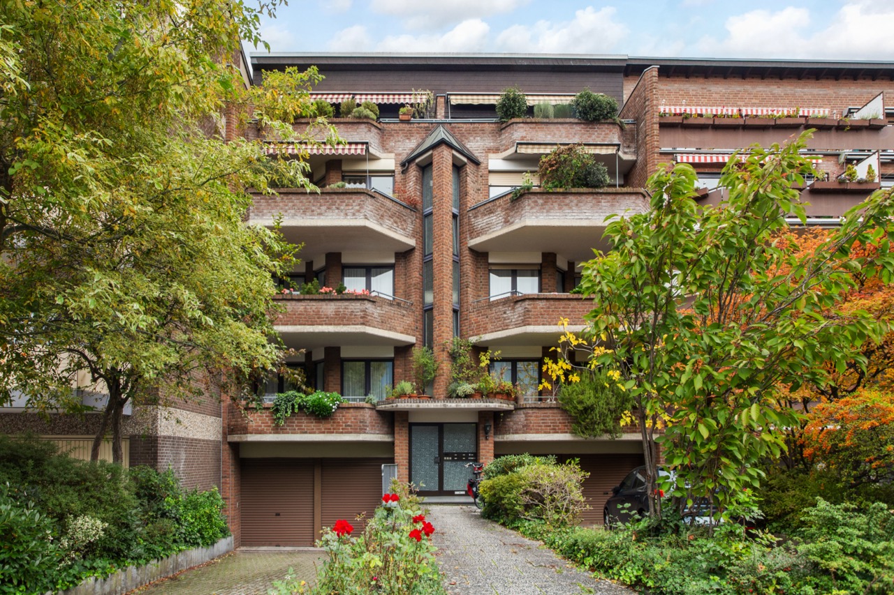 Mehrstöckiges Wohnhaus mit Balkonen, vielen Pflanzen und Bäumen im Vordergrund.