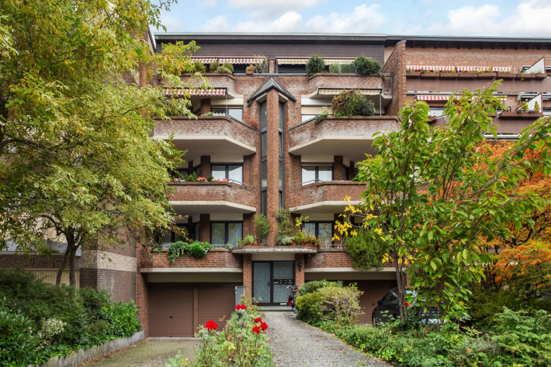 Mehrstöckiges Wohnhaus mit Balkonen, vielen Pflanzen und Bäumen im Vordergrund.