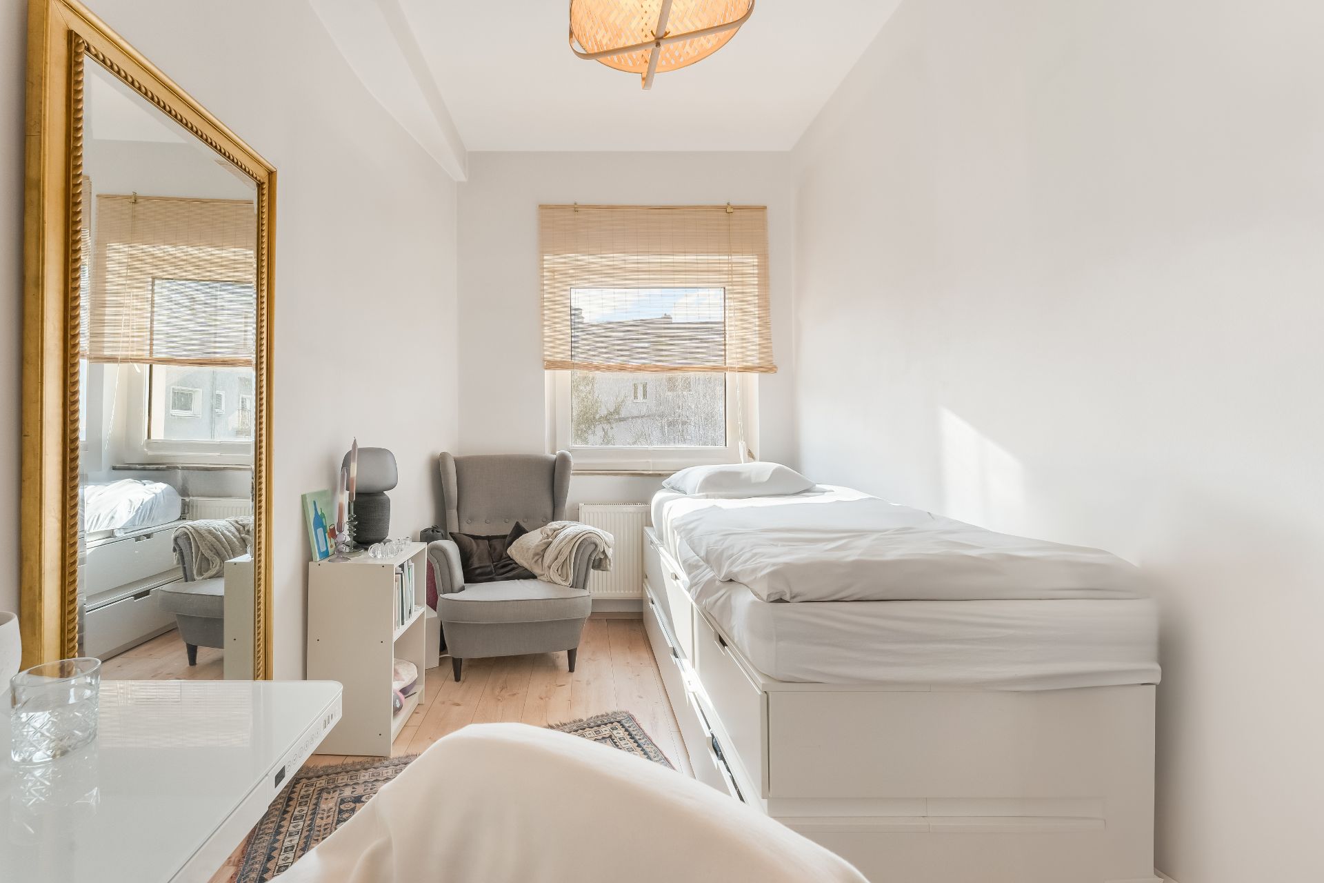 Helles Schlafzimmer mit Einzelbett, Sessel, Spiegel und Fenster mit Jalousie.