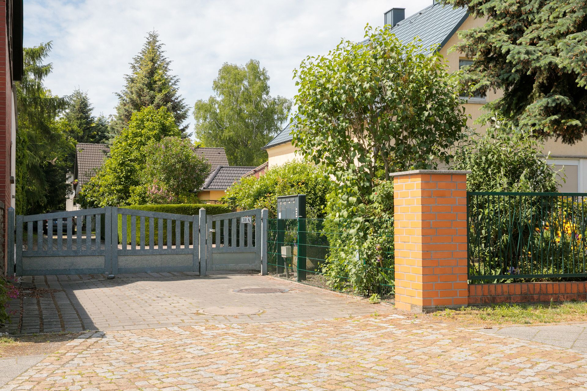 Einfahrt mit grauem Tor, Ziegelmauer und viel Grün in einem Wohngebiet.