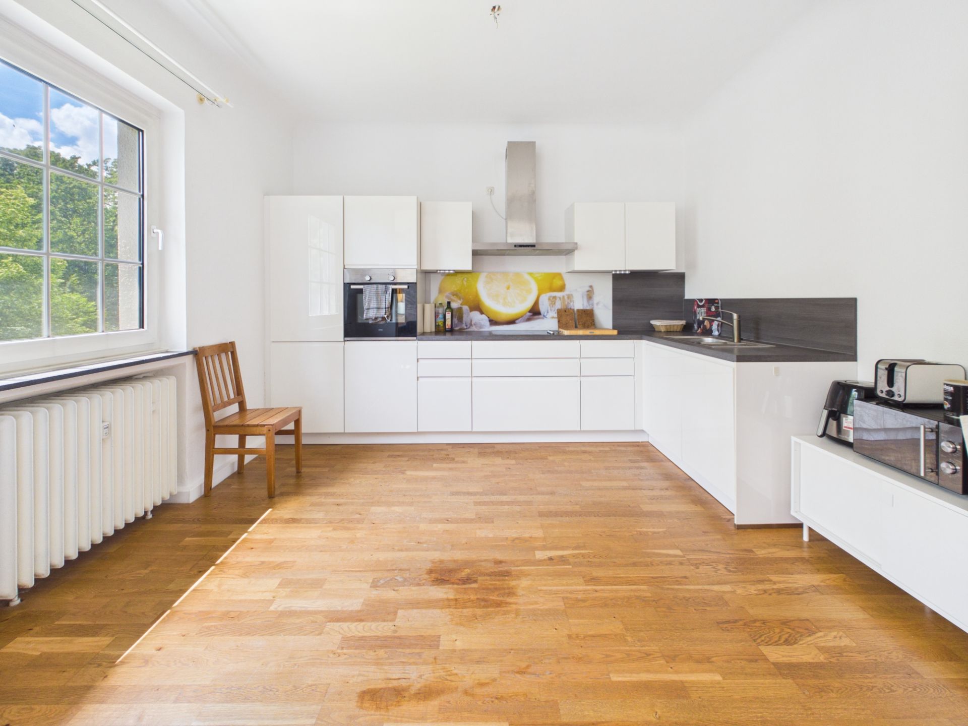 Moderne weiße Küche mit Holzfußboden, Fenster, Stuhl und Küchengeräten auf der Arbeitsplatte.