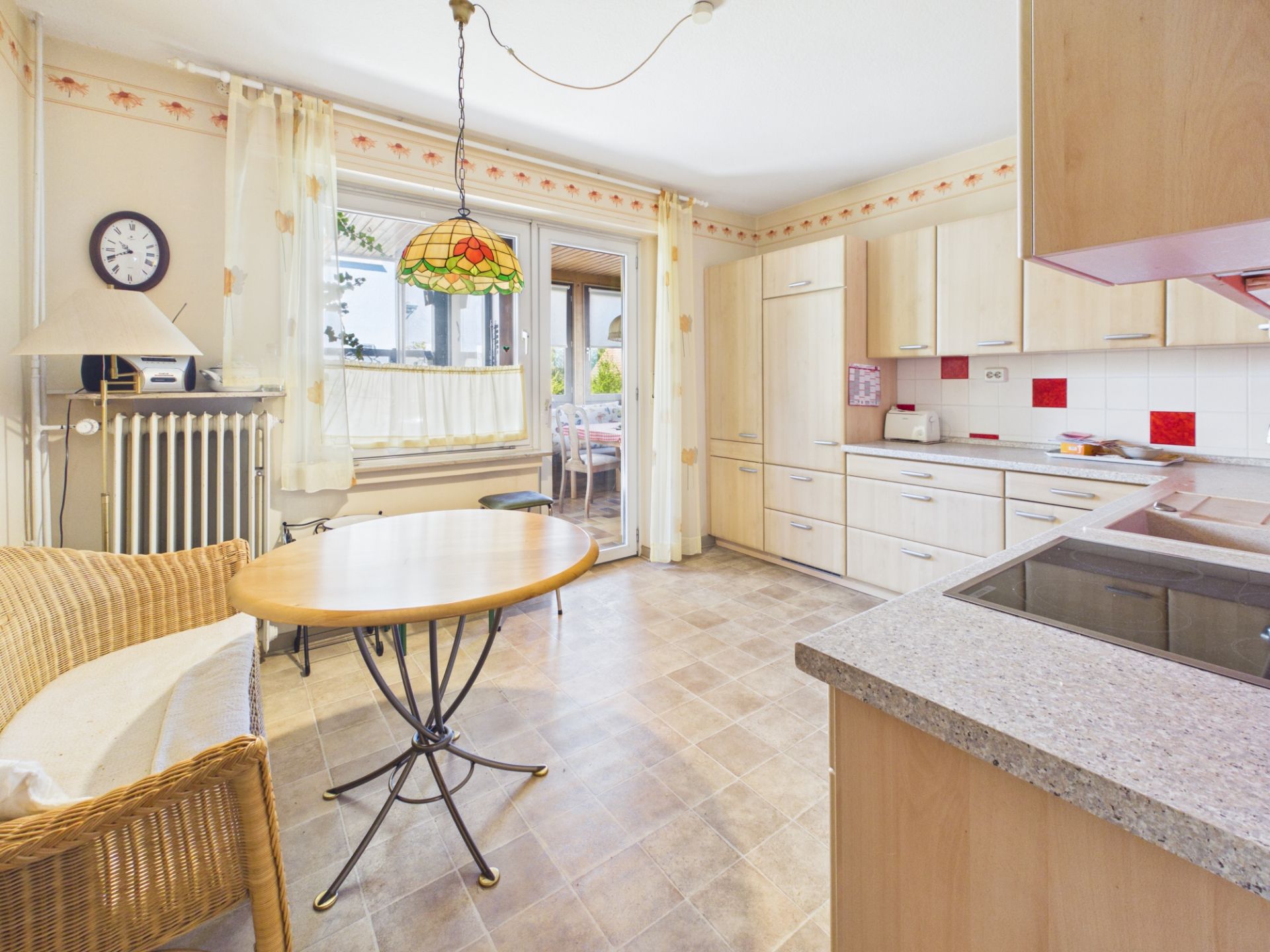 Helle Küche mit Holzmöbeln, rundem Tisch, Korbstuhl und Zugang zu einem Balkon.