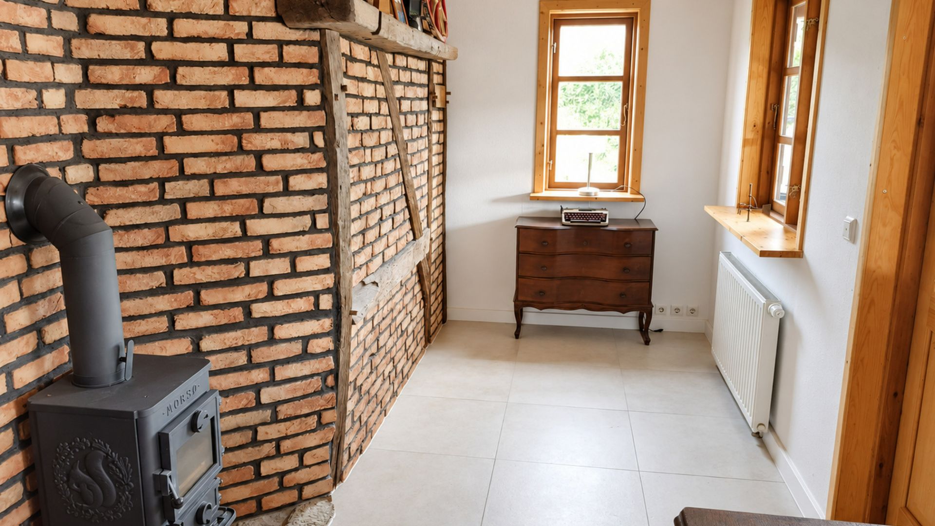 Zimmer mit Backsteinwand, Holzofen, Kommode und zwei Fenstern mit Holzrahmen.